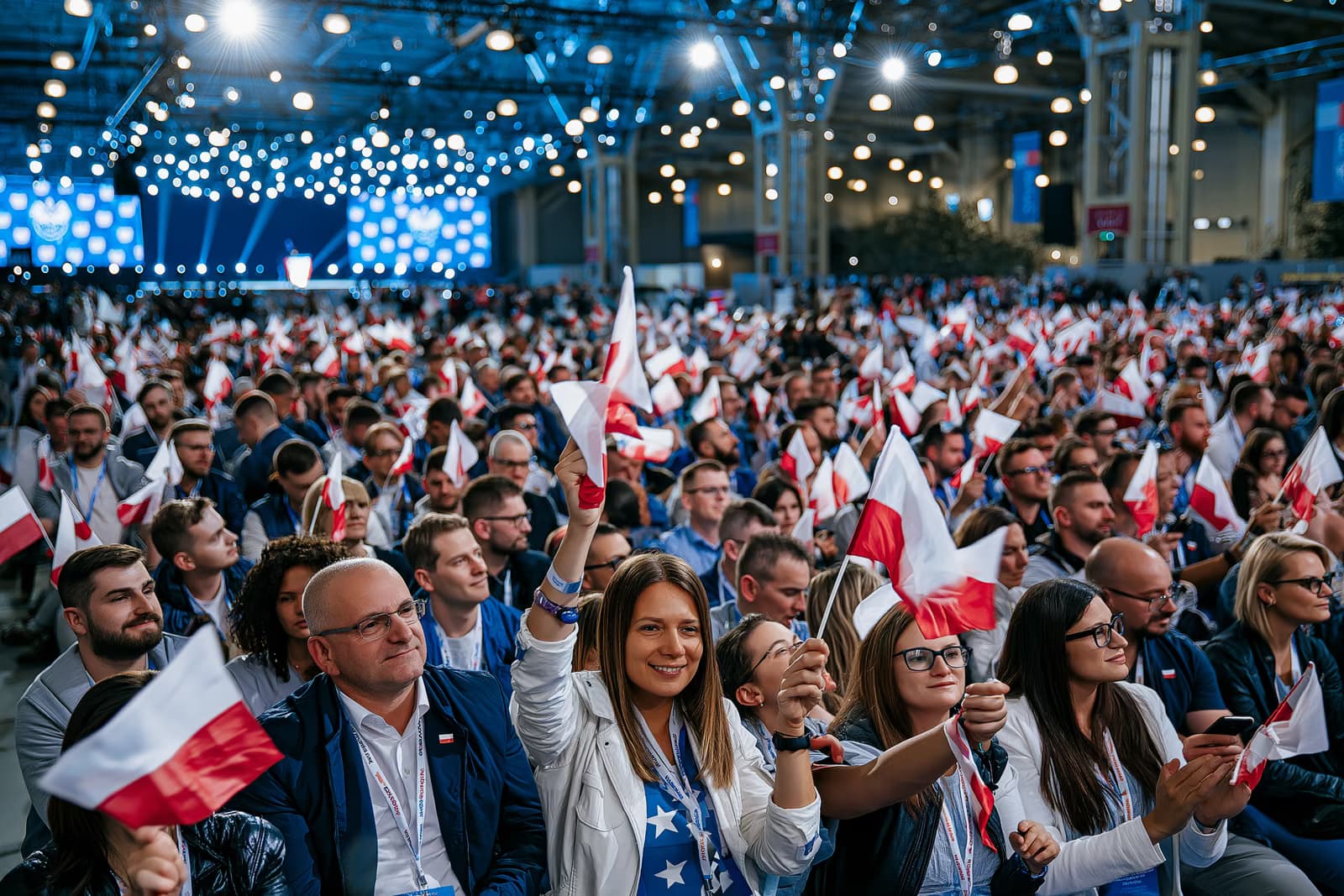 A packed exhibition hall crowd waving small flags under harsh event lighting at the AfD Magdeburg campaign launch, with a stage in the distance.