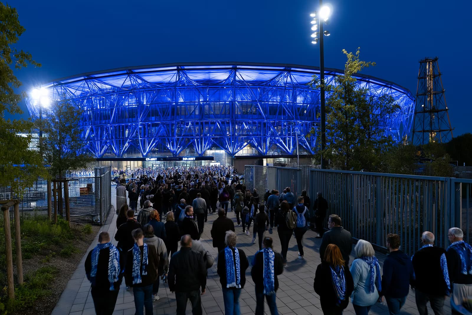 A football stadium in the Ruhr area glows royal blue at night as fans in scarves stream toward the gates after the Gosens Schalke move