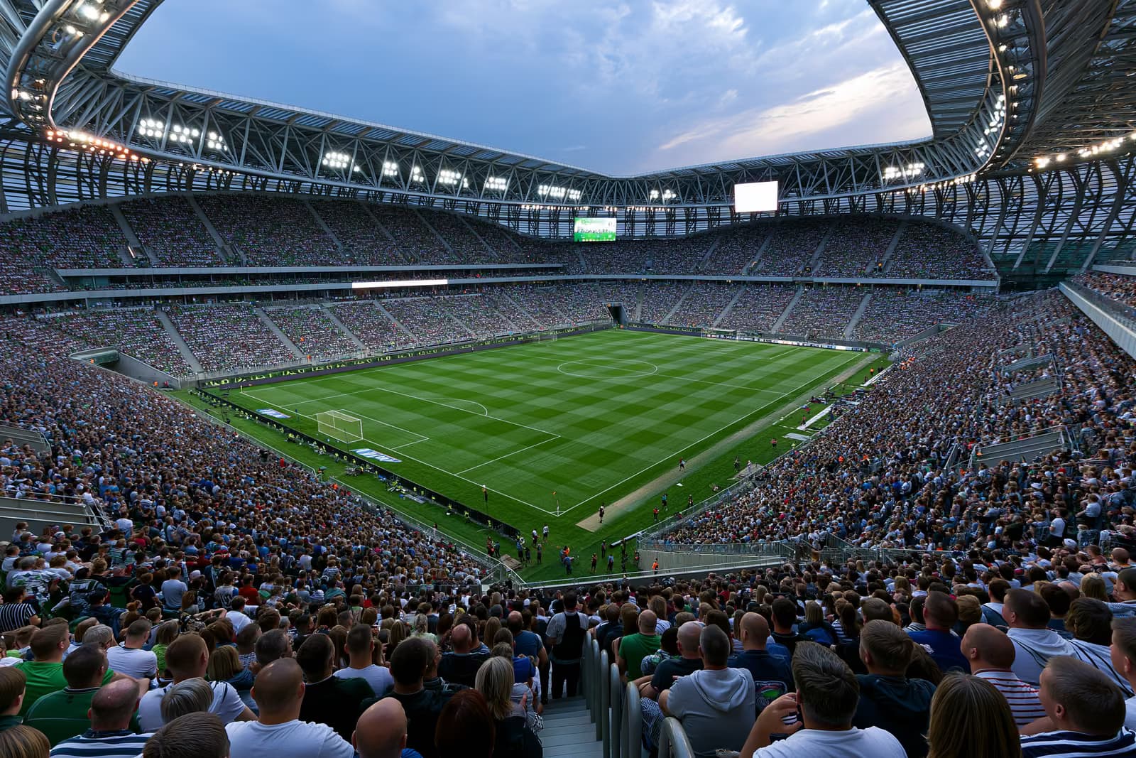 A large football stadium filled with fans under floodlights on a summer evening.