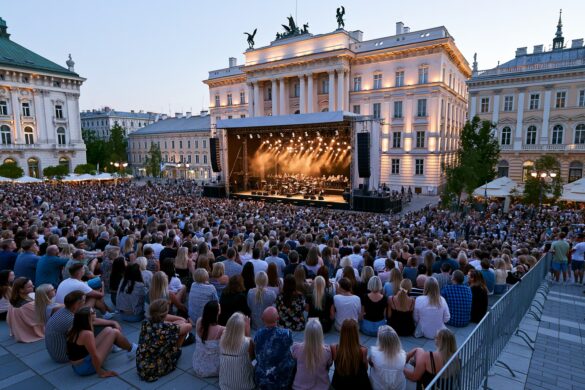 A crowd gathers at dusk on a historic Berlin square where an open-air orchestra stage is lit against grand facades.