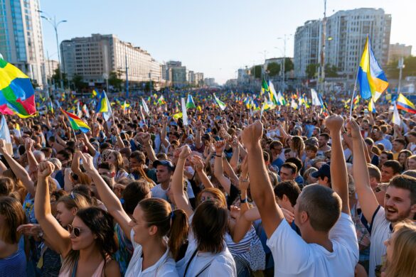 A huge crowd fills a city street under bright summer sky waving colorful flags.
