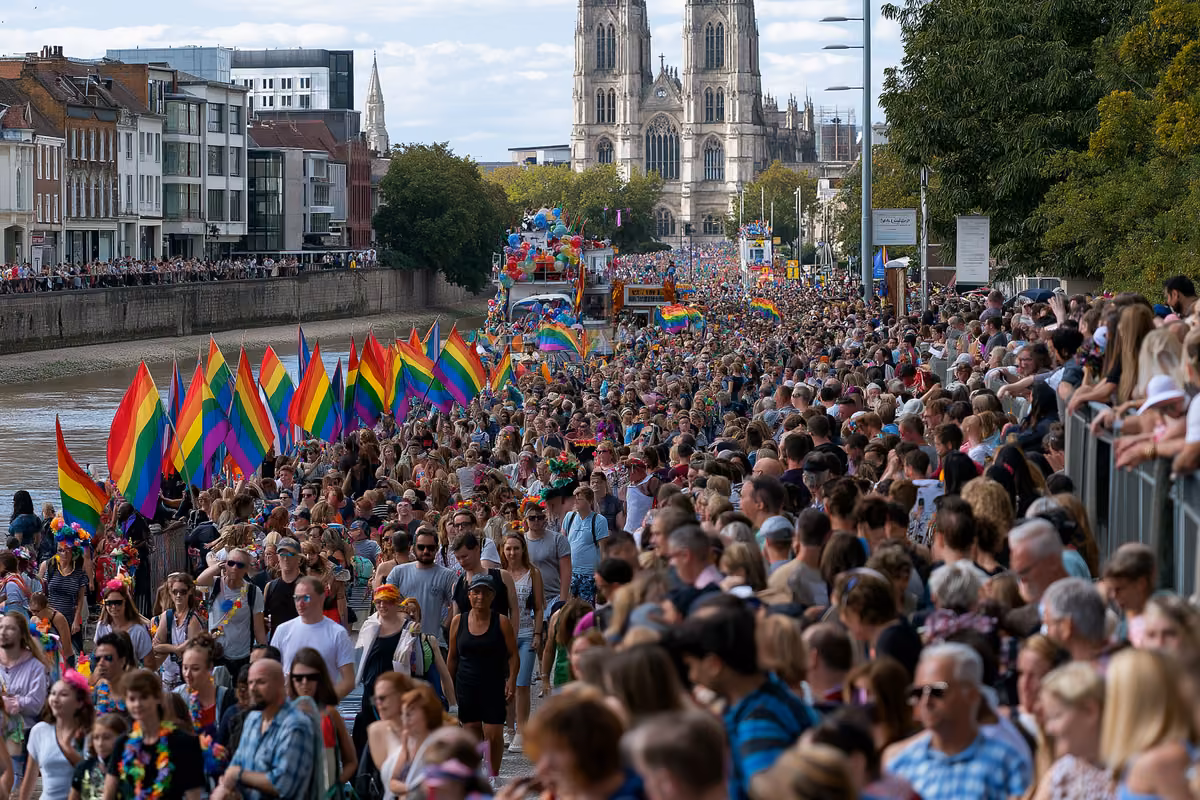 Rainbow flags and decorated floats moving through a crowded riverside street.