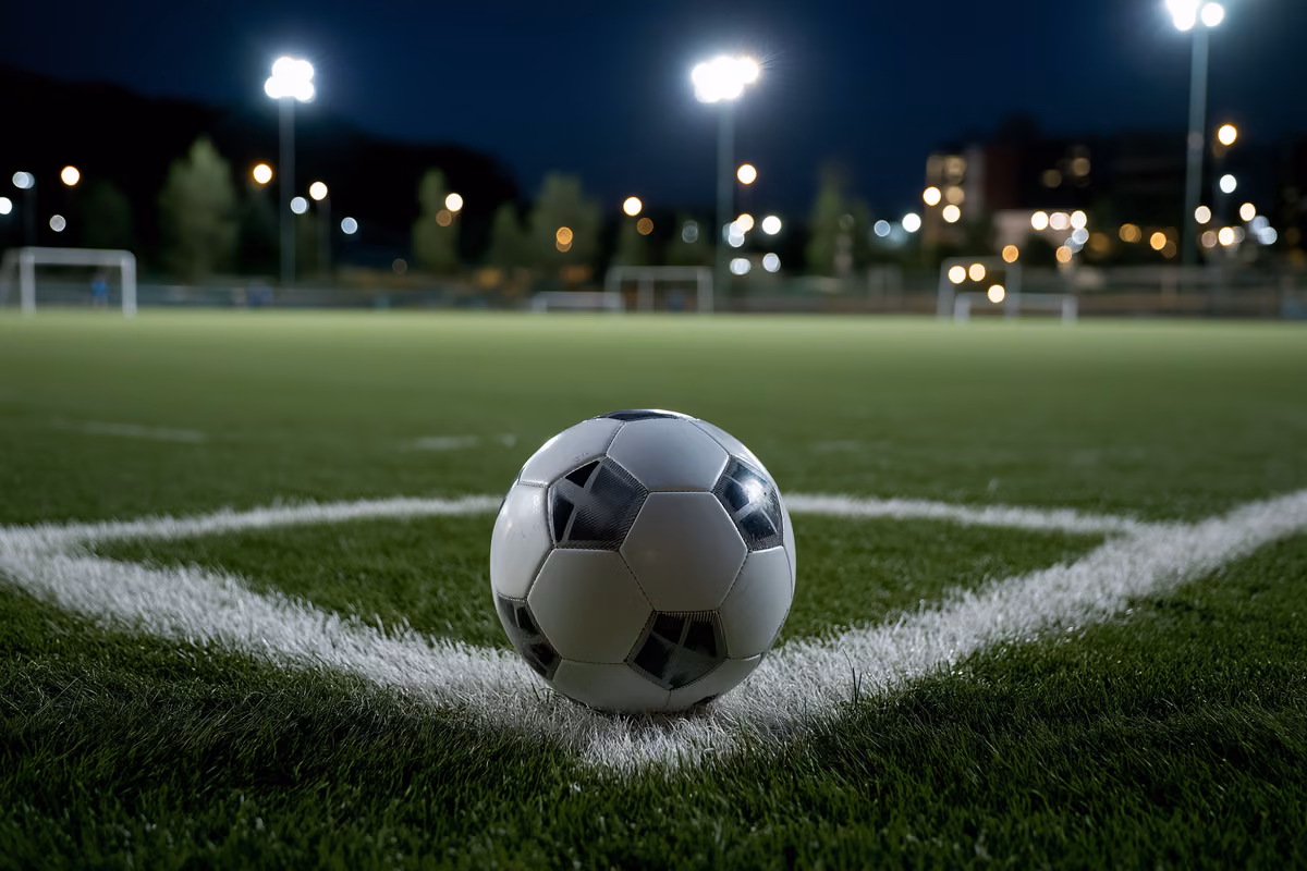 A lone football resting on the penalty spot of a floodlit pitch.