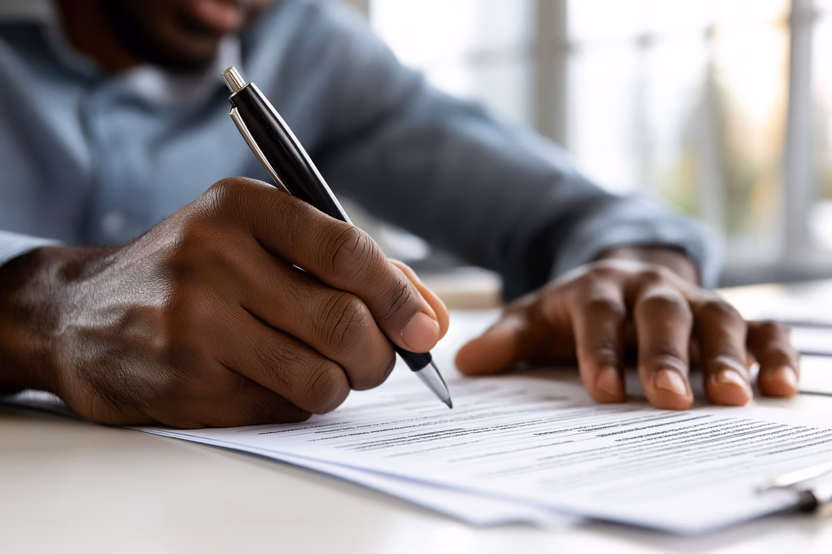 A person filling out paperwork at a desk with a pen and official forms.